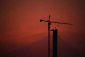 Silhouette of a tall construction crane against a vibrant red sunset sky, with cables from a bridge converging towards the base of the crane.