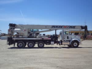 A boom truck with an extended boom arm parked on a gravel lot. The truck has a white cab and a company name "MAXIM" written on the side of the boom. The vehicle is equipped with stabilizing outriggers and has multiple axles to support its heavy lifting capabilities. The clear blue sky and an industrial area are visible in the background.