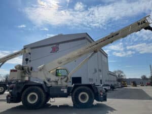A large white rough terrain crane parked outside a Maxim Crane Works building. The crane's extended arm reaches high, showcasing its lifting capacity. The sky is clear with a few scattered clouds, and additional construction equipment and vehicles are visible in the background.