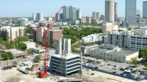 Aerial view of a construction site in an urban area with a prominent red Maxim Crane crawler crane in operation. The crane is being used to assemble a multi-story building. Surrounding the construction site are various residential and commercial buildings. In the background, the city skyline features tall skyscrapers and dense urban development.