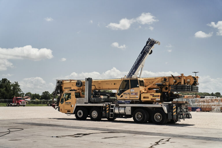 A yellow Maxim Crane hydraulic truck crane is positioned on a wide concrete lot with a backdrop of a partly cloudy sky. The crane has a telescoping boom that is partially extended and rests on the truck's chassis. The truck features multiple axles with large black tires and various hydraulics and support structures are visible along its sides. In the distance, there are other construction equipment and vehicles along with greenery and trees. Crane was manufactured by Grove.
