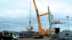 A yellow all-terrain Maxim Crane is lifting a large white cylindrical object at a construction or industrial site. Several workers in safety vests and helmets are present around the site, along with trucks and other machinery. The setting appears to be a port or dock, indicated by the “PORT STOCKTON” signage visible in the background. The sky is overcast, contributing to the industrial atmosphere of the scene.