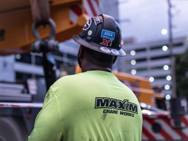Maxim Crane Works worker in safety gear operating crane at construction site.