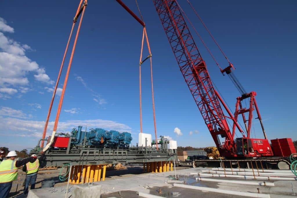 Crane lifts heavy machinery at construction site under clear blue skies.