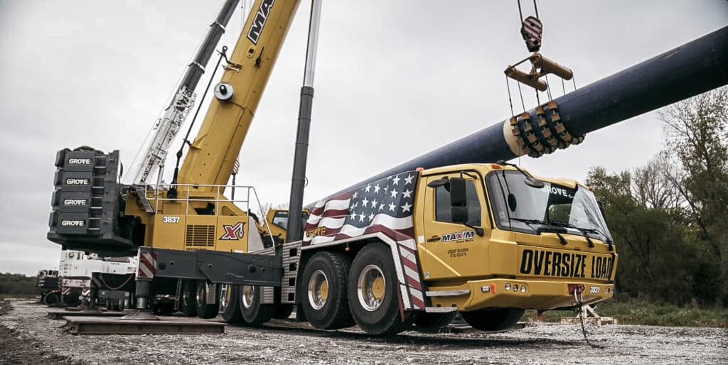 Crane lifting large pipe at construction site with American flag design.