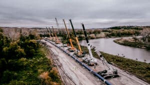Cranes operating at a riverbank construction site amid natural scenery.