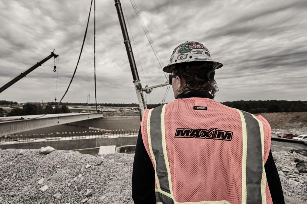 Construction worker overseeing bridge project with cranes under a cloudy sky.