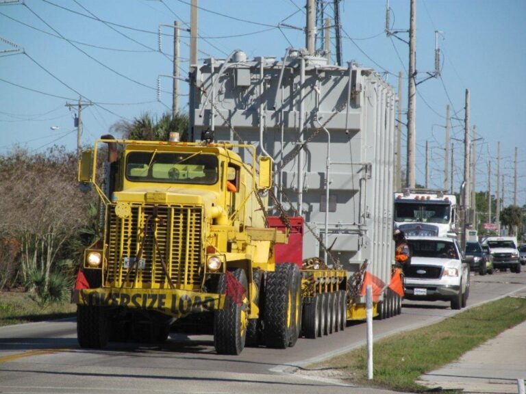 Oversized yellow truck carrying heavy equipment through crowded city streets.