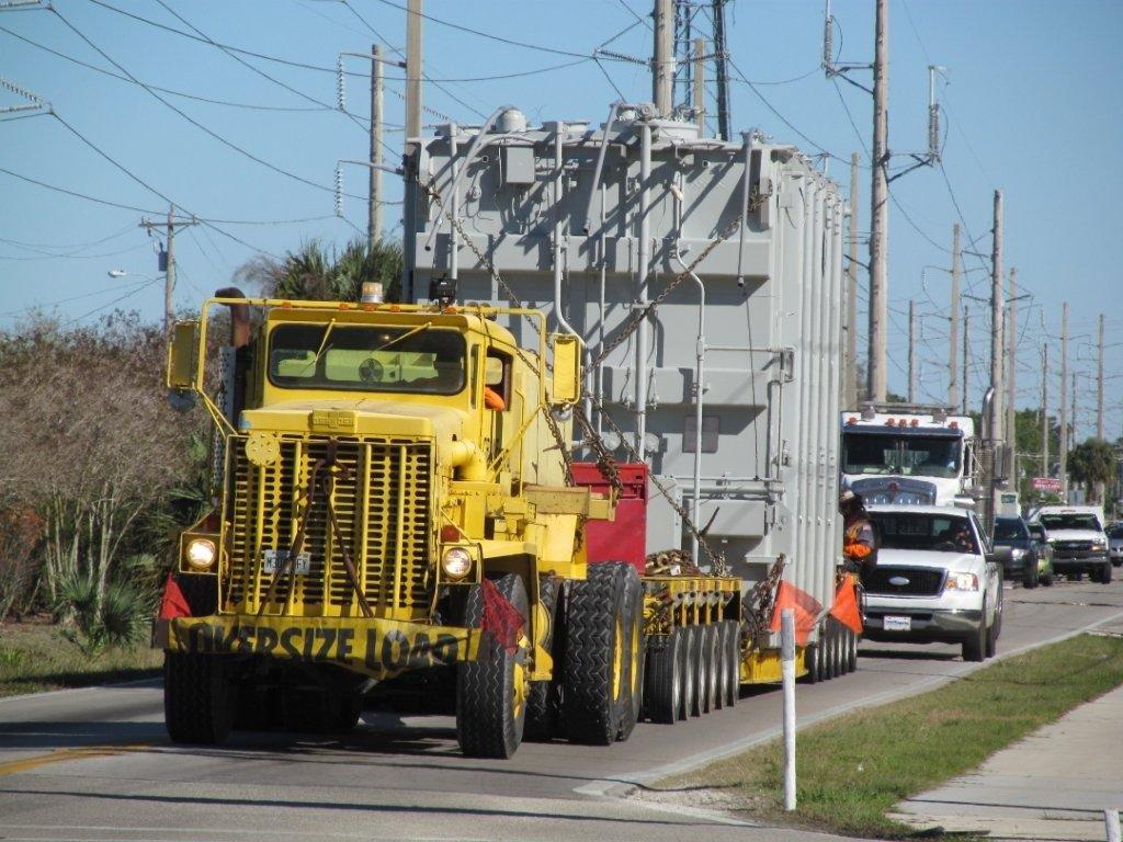 Oversized yellow truck carrying heavy equipment through crowded city streets.