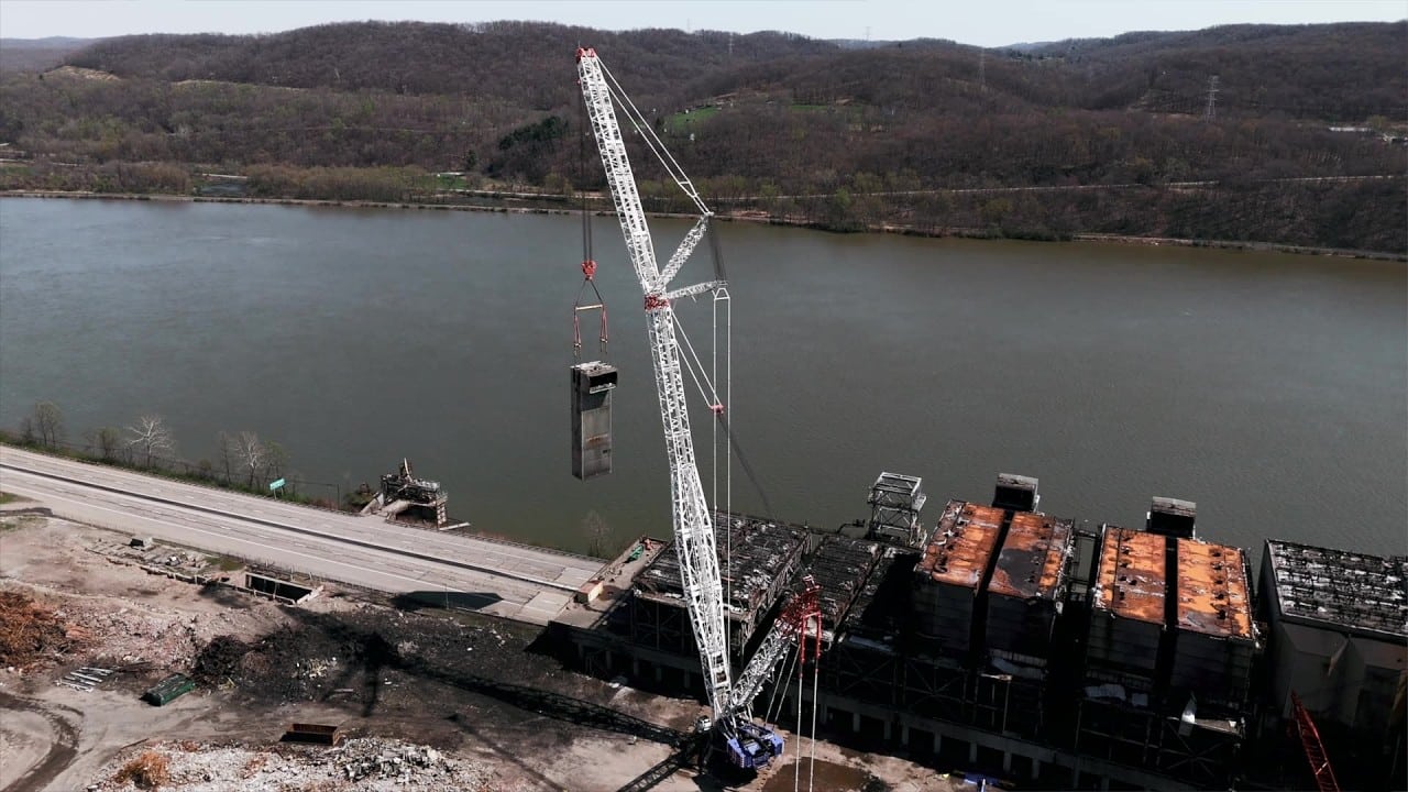 Crane lifting container over water at construction site with hills in background.