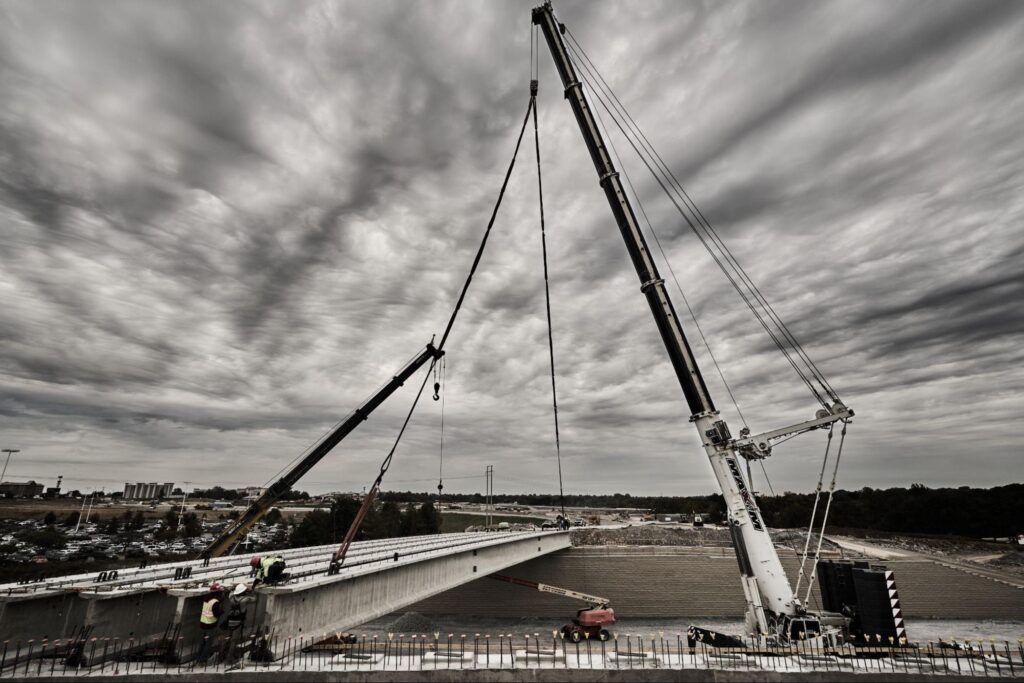 Construction workers operate cranes to build a new bridge against a dramatic sky.