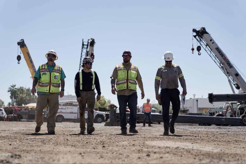 Construction workers walking on site with machinery in the background under clear skies.